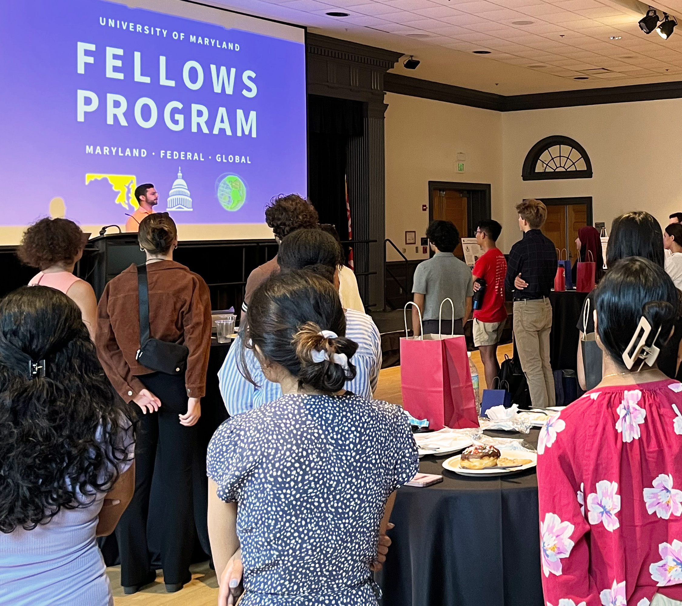 Many diverse new Fellows students in a ballroom listning to Jonathan Lee speaking on stage in front of the UMD Fellows Program Logo